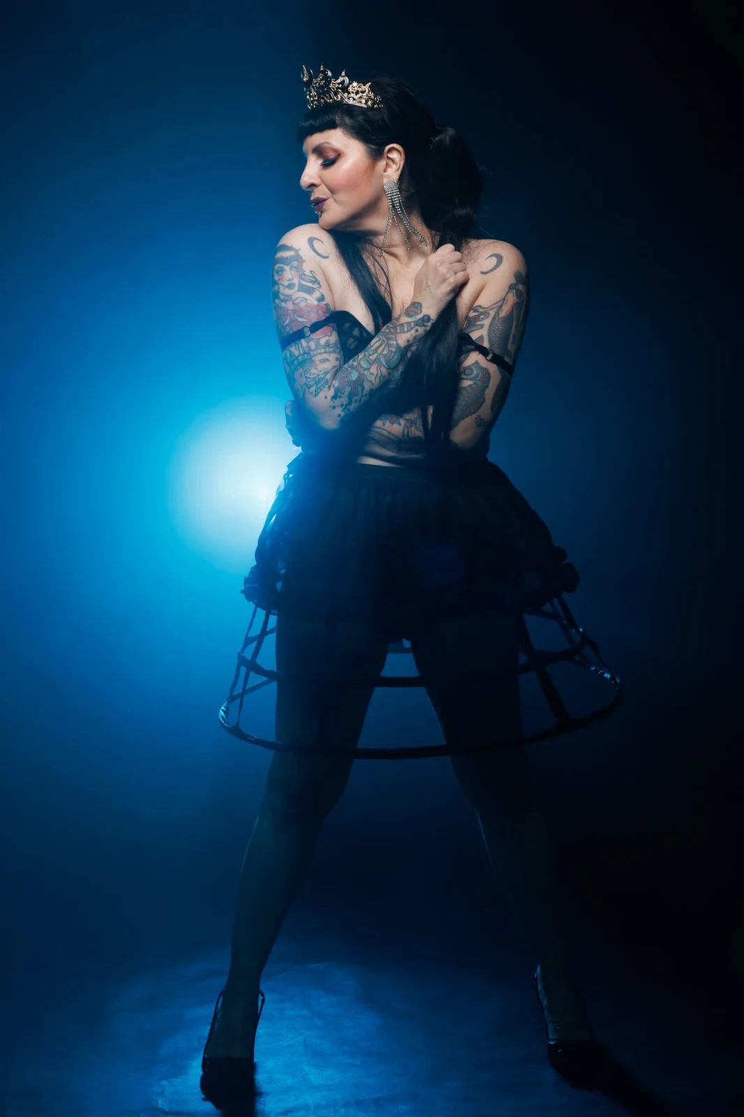 Anaïs Lune turning her face to the side in a crown and black hoop skirt against blue light