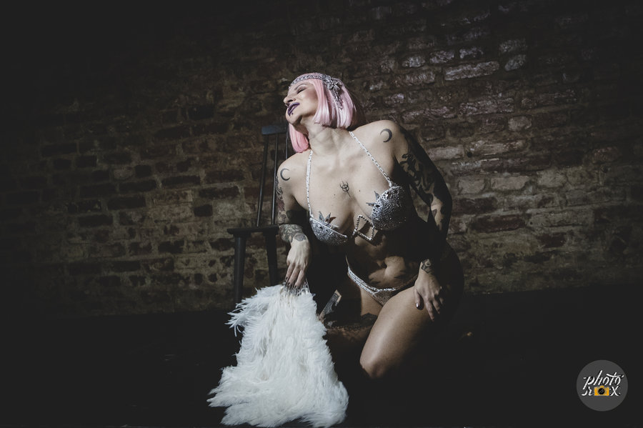 Anaïs Lune in a blonde wig kneeling on stage holding a white feather boa