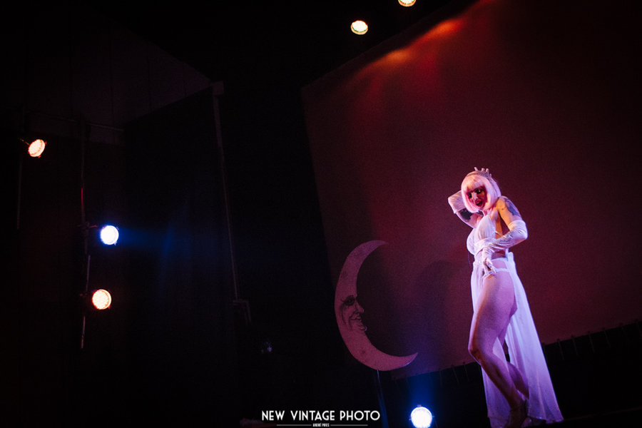 Anaïs Lune in a white flowing gown on a dark stage near a crescent moon prop