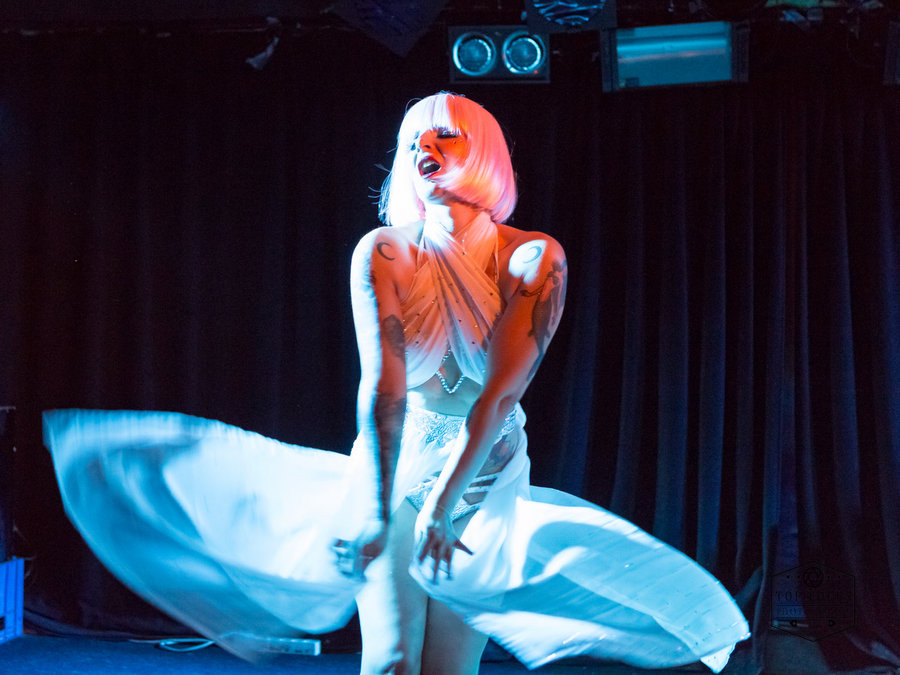 Anaïs Lune in a white dress catching air against a blue-lit curtain backdrop