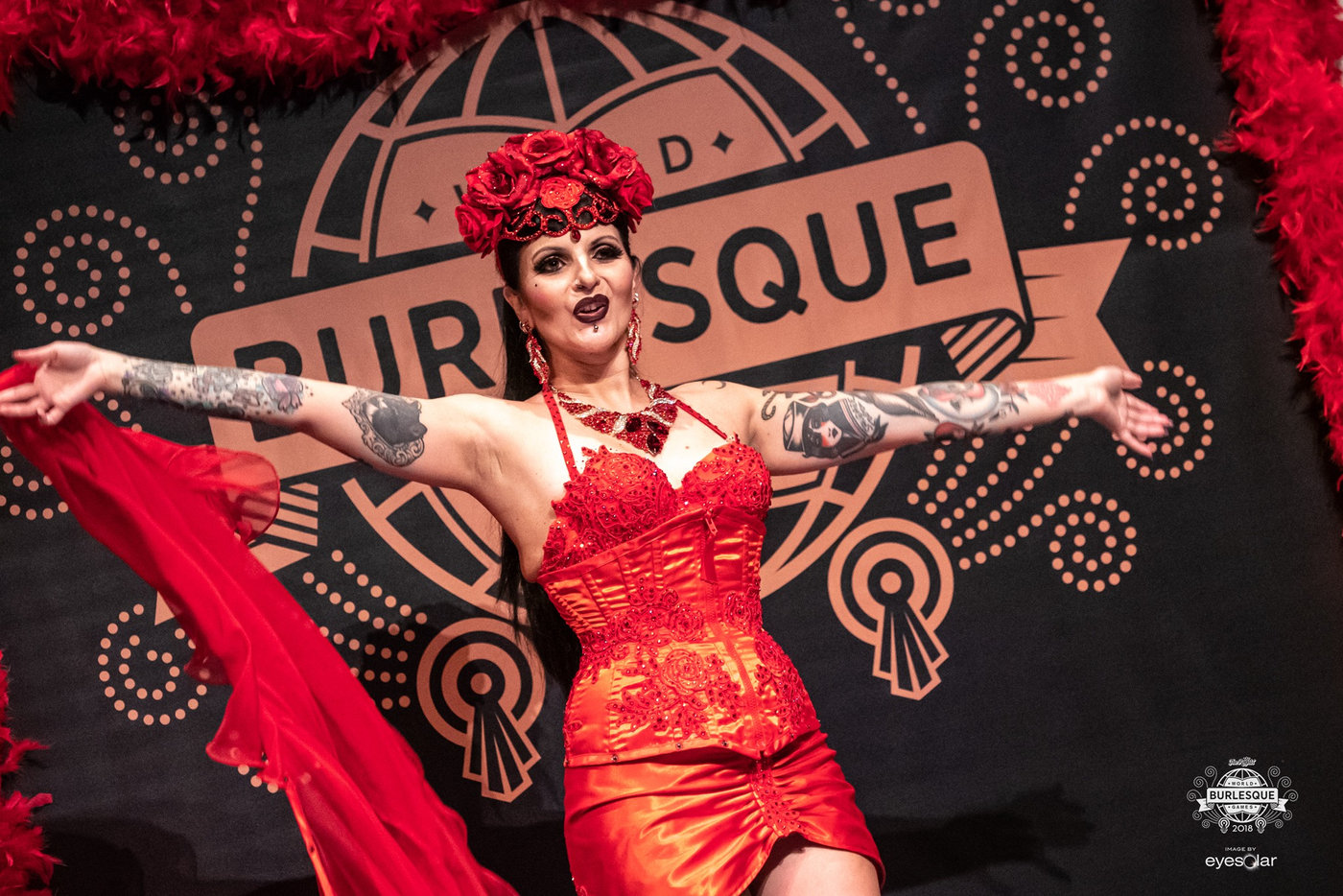 Anaïs Lune performing live on stage in a red dress with feather boas, in front of a burlesque marquee